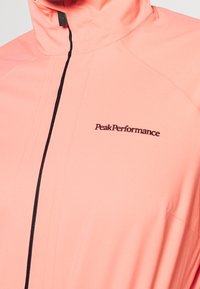 Veste légère à fermeture éclair de couleur corail vif. Comprend un col montant et un logo noir "PeakPerformance" sur la poitrine. Texture lisse.