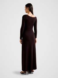 Long-sleeve, dark purple dress with a boat neckline and fitted silhouette, featuring a smooth texture. Worn with black heeled sandals.