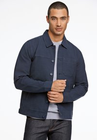 Marineblauwe jas met knoopsluiting, een borstzak en een gladde textuur. Gecombineerd met een grijze shirt met ronde hals eronder.