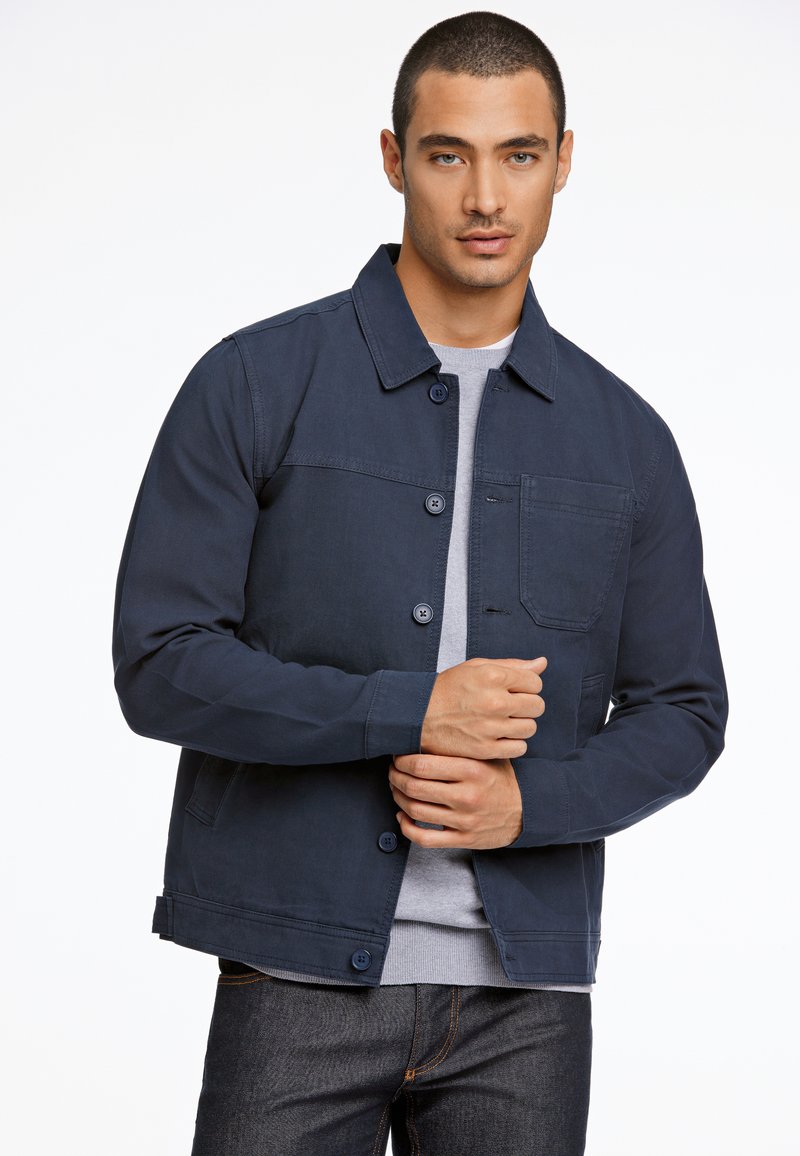 Marineblauwe jas met knoopsluiting, een borstzak en een gladde textuur. Gecombineerd met een grijze shirt met ronde hals eronder.