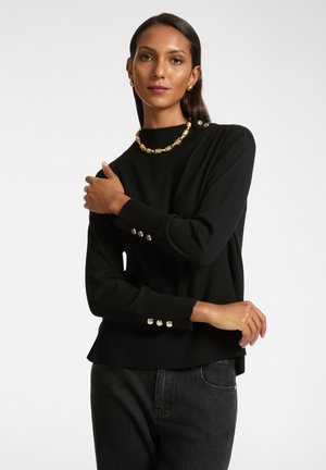 Maglione nero con collo alto, polsini a costine e bottoni decorati sulle maniche. Abbinato a jeans scuri e collana statement dorata.