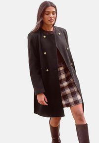 Cappotto nero doppiopetto con bottoni dorati, design aderente, indossato sopra una mini gonna a quadri. Stivali lucidi marroni fino al ginocchio completano l'outfit.