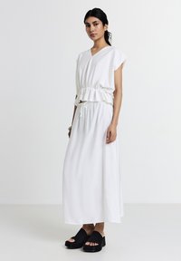 Femme portant un top péplum sans manches blanc avec une jupe longue assortie et des sandales à platforme noires ouvertes, se tenant devant un fond uni.