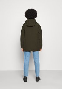 Parka verde con cappuccio, dal taglio dritto e texture. Abbinata a jeans azzurri chiari e scarpe nere, vista da dietro.