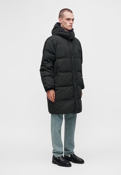 Homme debout portant un long manteau matelassé noir avec capuche, un jean bleu clair et des chaussures en cuir noir sur un fond blanc uni.