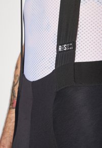 Cuissard de cyclisme noir avec partie supérieure en tissu en maille, arborant un label logo et des bretelles réglables. Texture textile lisse avec des panneaux contrastants.