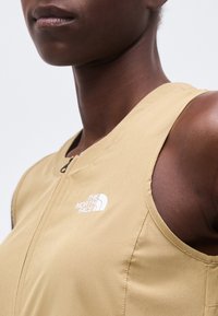 Κοντινό πλάνο ενός ατόμου που φοράει ένα μπεζ αμάνικο γιλέκο με φερμουάρ και το λογότυπο The North Face στο στήθος.