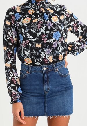 Vrouw draagt een zwarte bloemenblouse met lange mouwen, een geplooide halslijn en manchetten, gecombineerd met een blauwe denim minirok.
