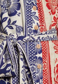 Vestido de algodón con estampados florales en azul y rojo sobre un fondo crema; incluye botones de madera y un cinturón con lazo. Tejido texturizado.
