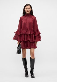 Burgunderkjole med klokkesløfter og lagdelte volanger, laget av gjennomsiktig stoff. Båret med svarte høye støvler og en svart naglet håndveske.