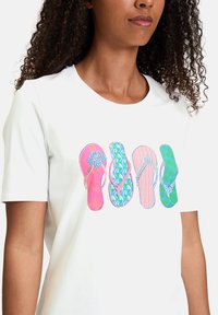 T-shirt blanc avec un graphique de quatre tongs en rose, vert et bleu, présentant divers motifs, notamment des motifs floraux et rayés.
