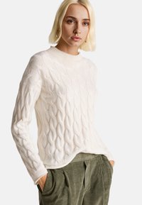 Krémový svetr s pleteným vzorem, kulatým výstřihem a dlouhými rukávy, kombinovaný se zelenými kordové kalhotami.