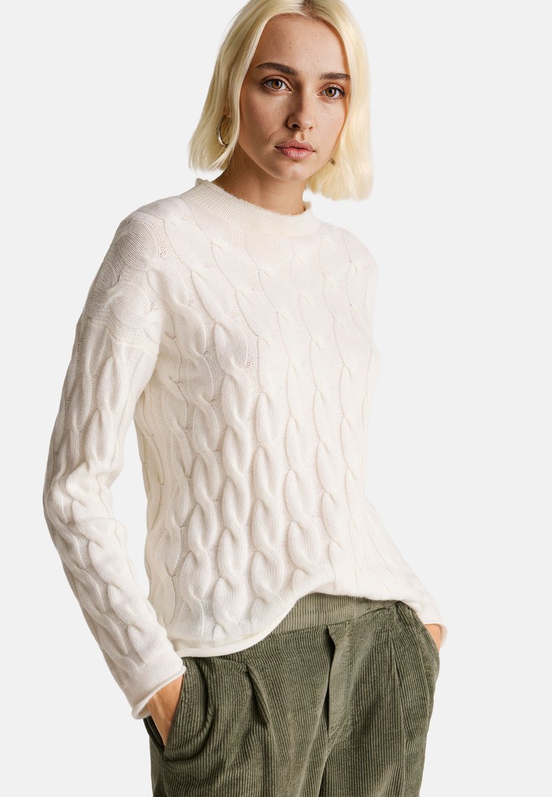 Krémový svetr s pleteným vzorem, kulatým výstřihem a dlouhými rukávy, kombinovaný se zelenými kordové kalhotami.