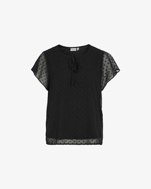 Svart bluse med korte gjennomsiktige ermer, rund hals, en knytedetalj foran, og teksturert prikkemønster over hele.