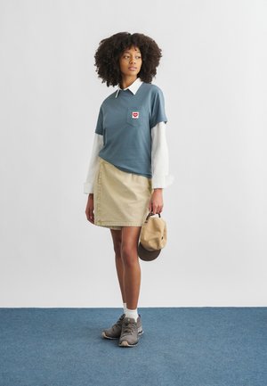 Młoda kobieta o kręconych włosach, ubrana w nałożone na siebie niebieskie i białe topy, beżową spódnicę, szare trampki, trzymająca beżową czapkę, stojąca na niebieskim dywanie.