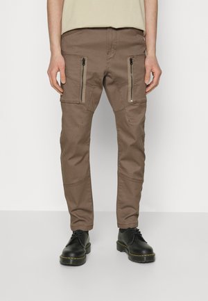 Braune Slim-Fit-Cargohosen mit vorderen Reißverschlusstaschen, kombiniert mit schwarzen Schnürstiefeln, gezeigt von der Taille bis zu den Füßen vor einem schlichten Hintergrund.