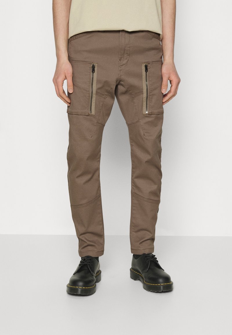 Pantalon cargo marron slim-fit avec poches zippées à l'avant, porté avec des bottes noires à lacets, présenté de la taille aux pieds sur un fond uni.