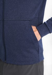 Giacca blu zip-up con una texture melangiata, dotata di tasche laterali e polsini aderenti. Il tessuto appare morbido e leggero.