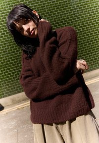 Brown fluffy sweater with ribbed hem, worn over light-colored corduroy pants. Background features green tiled wall.