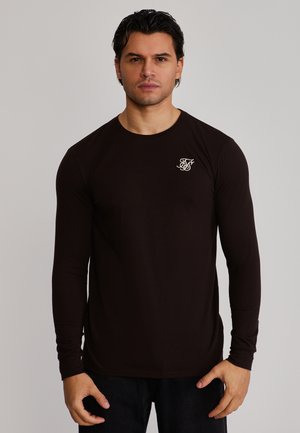 Uomo con capelli scuri che indossa una maglia nera a maniche lunghe con un piccolo logo bianco sul petto sinistro, in piedi davanti a uno sfondo semplice.