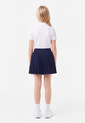 Pige stående med ryggen til iført en hvid kortærmet poloshirt, navyblåt plisseret skørt, hvide sokker og hvide sneakers.