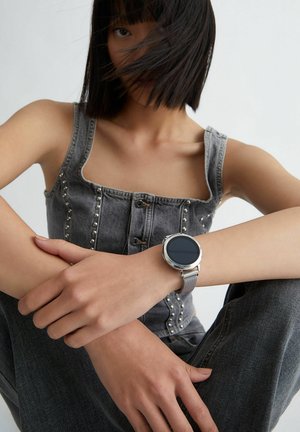 Montre intelligente argentée avec un cadran rond sur un poignet, affichage éteint. Portée avec un haut en denim gris avec des clous argentés et un décolleté carré.