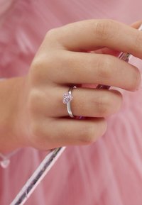 Silberner Ring mit einem herzförmigen rosa Edelstein, der an einer Hand getragen wird, die einen bunten Stock hält, vor einem sanften rosa Hintergrund.