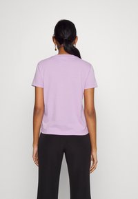 Kvinna med mörkt hår i hästsvans som bär en lavendelfärgad t-shirt med kort ärm och svarta byxor, stående vänd mot en enfärgad vit vägg.