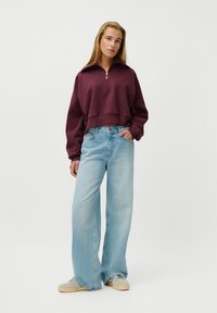 Young woman standing, wearing a cropped maroon zip-up sweatshirt, light blue wide-leg jeans, and light-colored sneakers on a plain white background.