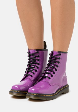 Bottes de combat en cuir verni violet avec lacets noirs, surpiqûres jaunes et semelle épaisse en caoutchouc, dotées d'une languette de traction bouclée à l'arrière.