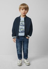 Marine bomberjack met witte strepen bij de manchetten, lichtblauw graphic t-shirt, donkerblauwe spijkerbroek en witte sneakers met grijze accenten.