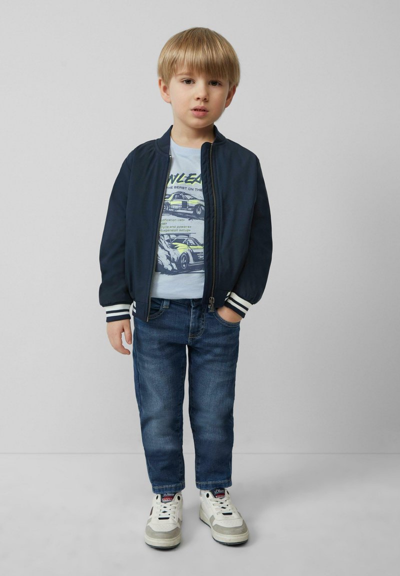 Marine bomberjack met witte strepen bij de manchetten, lichtblauw graphic t-shirt, donkerblauwe spijkerbroek en witte sneakers met grijze accenten.