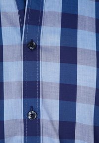 Koszula zapinana na guziki o niebiesko-granatowym wzorze w kratę, wykonana z lekkiego materiału, z czarnym guzikiem i białymi detalami przeszyć.