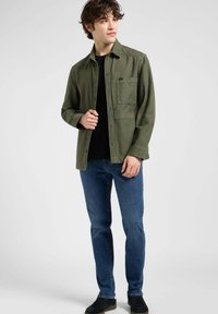 Chaqueta abotonada verde oliva con un bolsillo en el pecho, diseño con cuello, usada sobre una camisa negra y combinada con vaqueros azul oscuro. Zapatillas negras.