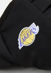 Juodas audinys su tekstūruota paviršiumi turi apvalią logotipą: geltoną krepšinio kamuolį su "LOS ANGELES LAKERS" purpuriniu tekstu.