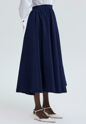 Žena v námornícky modrej midi sukni so zúženým pásom, bielej košeli s dlhým rukávom a bielych špicatých topánkach na podpätku, stojaca na svetlom pozadí.