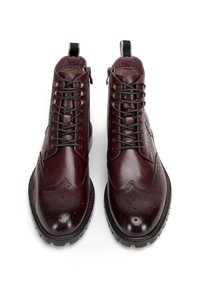 Burgundské kožené kotníkové boty s brogue detaily, kulatou špicí a kovovými očky. Obsahují černé tkaničky a strukturovanou podrážku.
