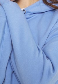 Osoba v měkké světle modré mikině s kapucí a stahovacími šňůrkami, se zkříženýma rukama přitisknutýma těsně k tělu, ukazující texturu a záhyby látky.