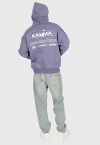 Person, der einen lila Hoodie mit weißem "KARMA"-Text und -Spruch auf dem Rücken trägt, kombiniert mit hellblauen Jeans und weißen Sneakers, schaut weg.