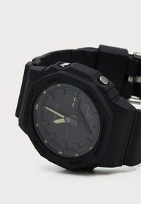 Ceas G-SHOCK din plastic negru, cu un cadran rotund, marcaje luminescente, bezel texturat și brățară din cauciuc. Dispune de butoane pe lateral.