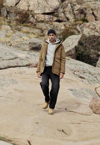 Jachetă izolată maro peste un hanorac gri, pantaloni negri și cizme bej. Fundalul prezintă un peisaj stâncos și vegetație rară.