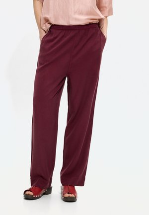 Kobieta w luźnych bordowych spodniach, pudrowo-różowej bluzce i czerwonych drewniakach z odkrytymi palcami ozdobionych srebrnymi ćwiekami, z rękami w kieszeniach, stojąca na białej podłodze.