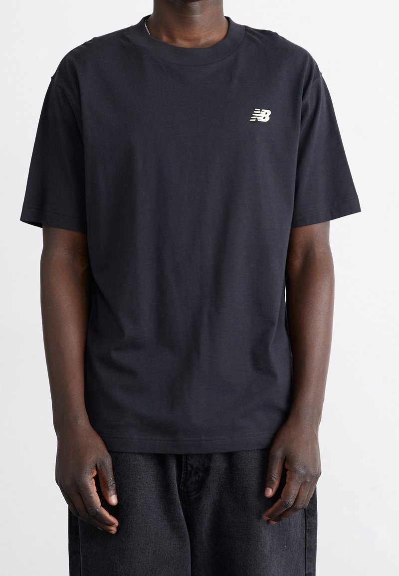 Camiseta de algodón negra con cuello redondo, que presenta un pequeño logo metálico dorado en el lado izquierdo del pecho. Mangas cortas y un ajuste relajado.
