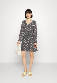 Vestido com padrão floral em tecido escuro; possui decote em V, mangas longas e faixa na cintura. Combinado com sandálias de salto amarelo-claro e uma bolsa branca.