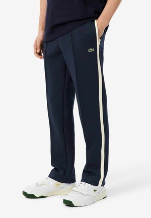 Persona che indossa pantaloni da ginnastica blu navy con strisce bianche ai lati e scarpe da ginnastica bianche con dettagli verdi, in piedi con una mano in tasca.