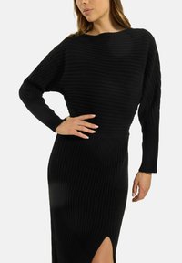 Schwarzes geripptes Strickkleid mit langen Ärmeln, Rundhalsausschnitt und seitlichem Schlitz für Bewegungsfreiheit. Die Textur ist weich und enganliegend.
