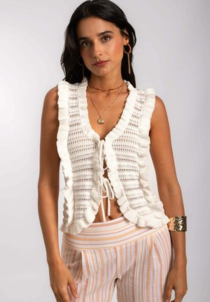 White knit vest with ruffled edges and a tie closure, featuring a loose, open design. Worn with striped pastel pants and gold accessories.