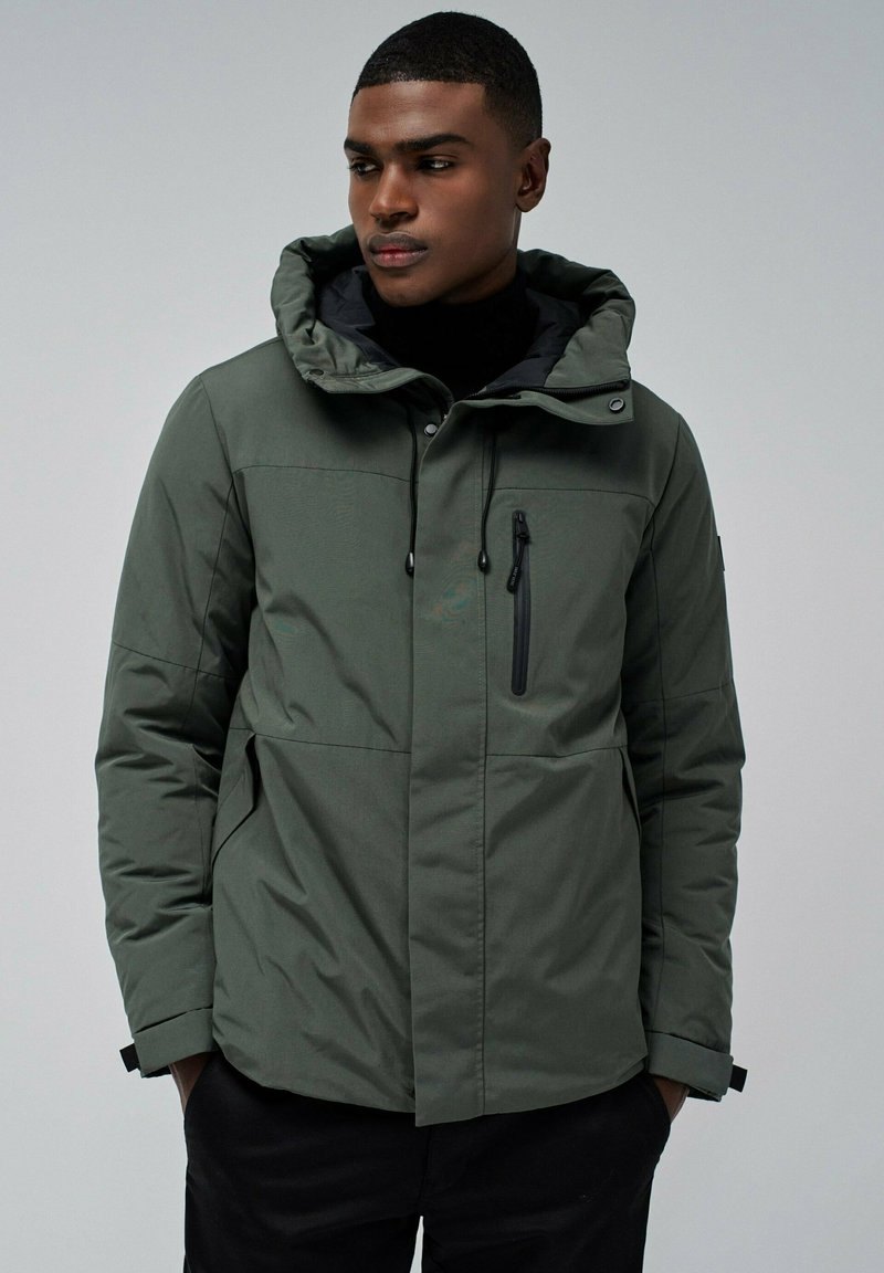 Hombre vistiendo una chaqueta de invierno con capucha de color verde oscuro, con un bolsillo con cremallera en el pecho y las manos en los bolsillos laterales, mirando hacia un lado.