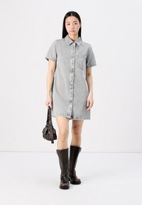 Vestido de ganga cinza de manga curta, com fecho de botão na frente, bolso no peito e um corte descontraído, combinado com botas castanhas até ao joelho e uma pequena bolsa.