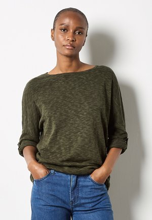 Haut olive décontracté à manches longues avec manches retroussées, en tissu texturé et décolleté large, associé à un jean en denim bleu.